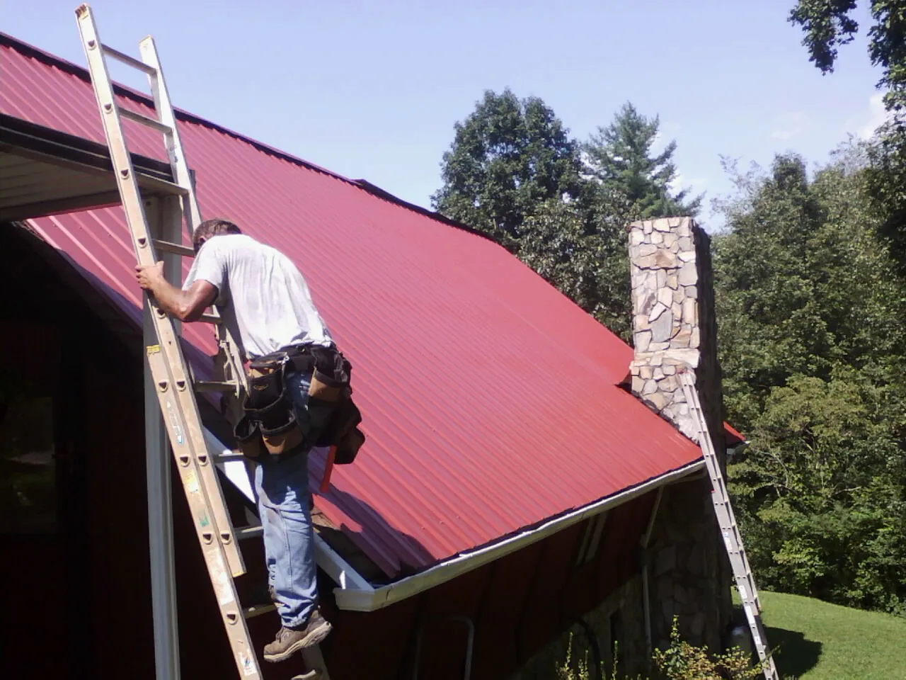 Roofer on a ladder beside a red metal roof and stone chimney — Soffit & Fascia Repair by Roofing Brothers in Berryville